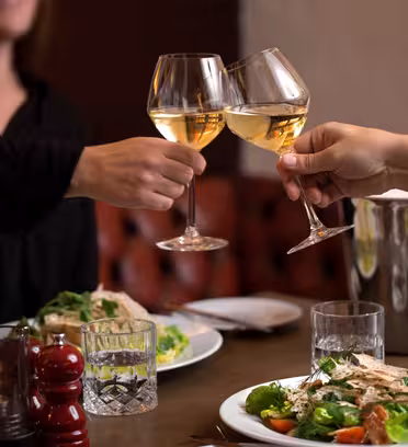To mennesker klirrende glas hvidvin over et spisebord med tallerkener med salat. Et stearinlys, vandglas og en vinkøler står på bordet. Atmosfæren er hyggelig og omgivelserne ser ud til at være en restaurant.