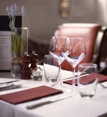 Et pænt dækket restaurantbord med to røde servietter, tallerkener, bestik, vinglas og vandglas. En menu, salt- og peberbøsser og en vase med lavendelblomster er placeret på den hvide dug. Røde stole er i baggrunden.