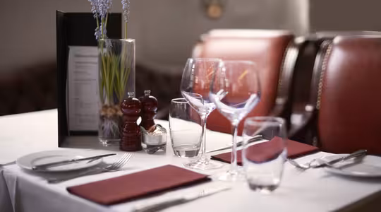 Et pænt dækket restaurantbord med to røde servietter, tallerkener, bestik, vinglas og vandglas. En menu, salt- og peberbøsser og en vase med lavendelblomster er placeret på den hvide dug. Røde stole er i baggrunden.