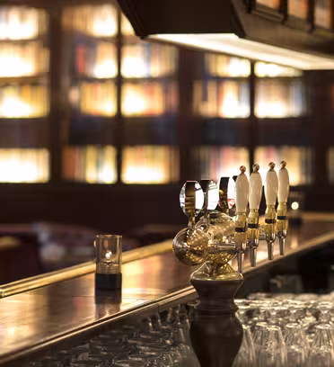 En svagt oplyst bar med en poleret trædisk, ølhaner og en række vendte glas. I baggrunden er der boghylder fyldt med bøger, hvilket skaber en hyggelig og indbydende stemning.