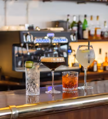 En bardisk i træ med fire drinks: en cocktail med agurkeskiver, en espresso martini, et whiskyglas med is og et hvidvinsglas. Bag dem ses en kaffemaskine og forskellige flasker.