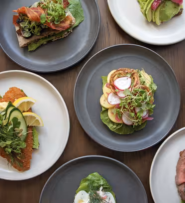 En række forskellige smørrebrød på tallerkener, med toppings som bacon, avocado, æg, rejer, grønt og radiser, arrangeret på et træbord. Hver sandwich er pyntet med friske krydderurter og citronbåde.