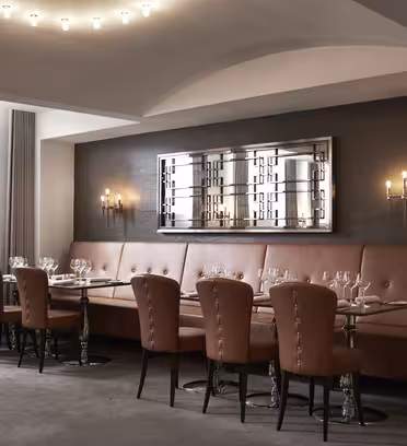 Elegant spiseplads med en lang brun banket langs en mørk væg, flankeret af læderstole og flere dækkede borde. Væggen har et geometrisk spejl og oplyste lampetter. Overhead, moderne runde lys giver belysning.
