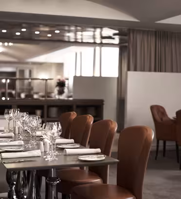Et moderne, elegant restaurantinteriør med pænt arrangerede borde med glas og servietter. Brune læderstole omgiver bordene, og bløde, neutrale toner fremhæver den rummelige og indbydende atmosfære.