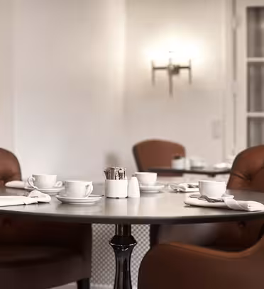 En hyggelig spiseplads med et rundt bord til fire. Bordet har hvide tekopper, underkopper og servietter. Brune polstrede stole omkranser bordet. En blød væglampe tilføjer varme til det neutrale rum.