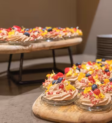 Små glaskrukker fyldt med cremede desserter er toppet med blåbær, ribs og spiselige blomster på to træbakker. Bakkerne placeres på en bordplade ved siden af en stak tallerkener.