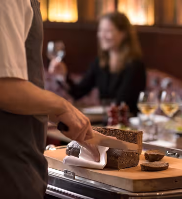 En person i et forklæde skærer et brød på et træskærebræt i en svagt oplyst restaurant. I baggrunden smiler en siddende person, der holder et vinglas. Glas hvidvin står på bordet.