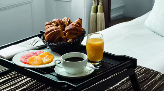 Morgenmad på sengen med en kurv med croissanter, en tallerken grapefrugt og appelsinskiver, et glas appelsinjuice og en kop kaffe på en bakke oven på en pænt redt seng. Rummet har bløde, neutrale toner.