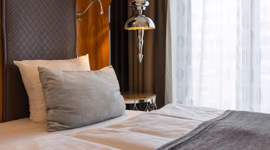 En pænt lavet seng med hvidt og gråt sengetøj er ved siden af et træpanel og en moderne metallisk lampe. Et stort vindue med rene hvide gardiner tillader blødt dagslys at oplyse rummet. Stemningen er rolig og indbydende.
